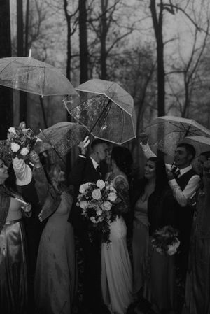 A wedding party stands together under clear umbrellas holding bouquets in a moody forest setting.