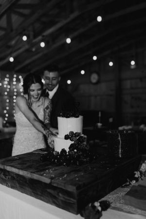 A couple cuts their elegant white wedding cake decorated with dark berries under string lights.