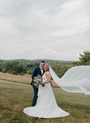 Bride's veil flows in the wind as couple poses on hilltop with scenic countryside view.