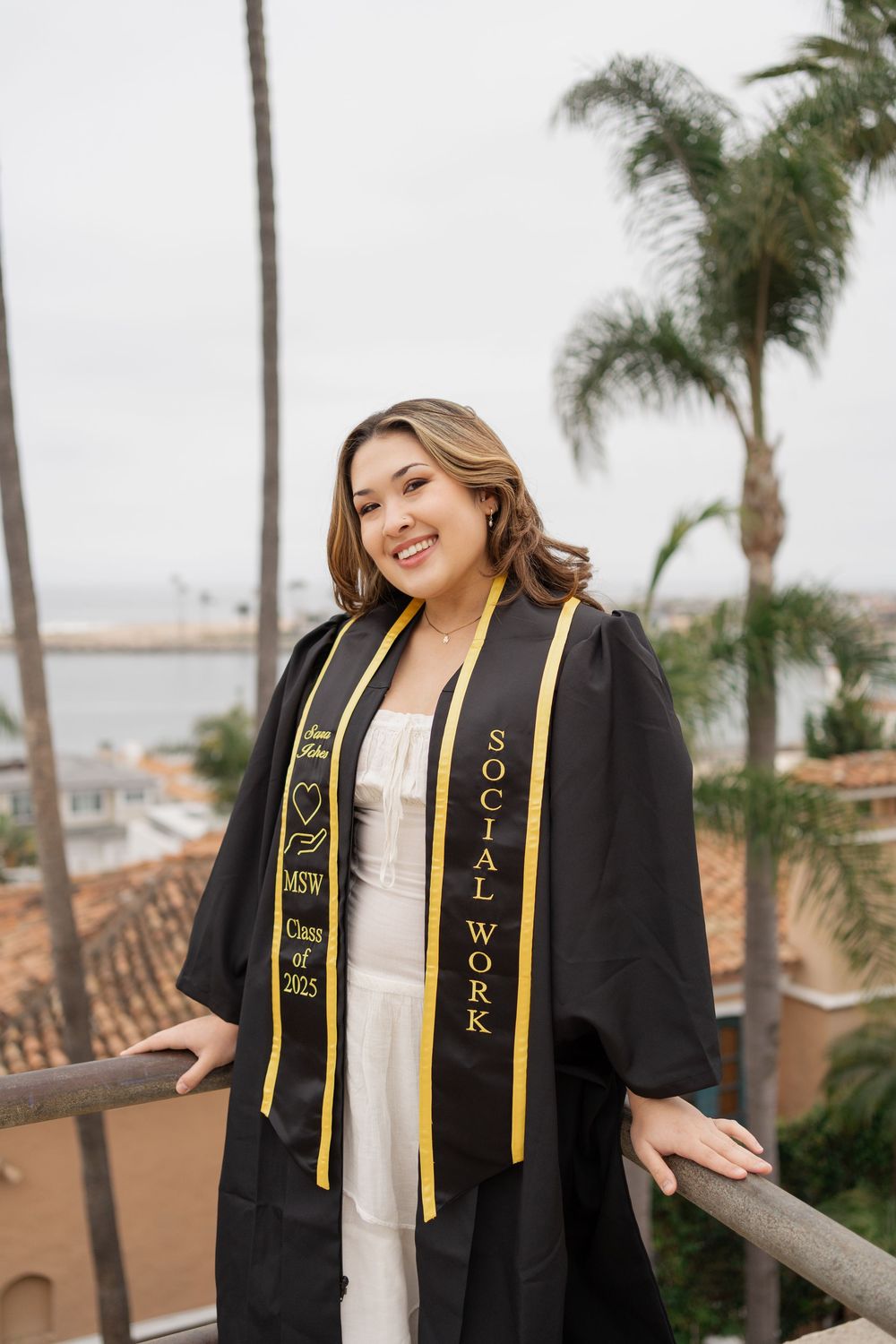 Smiling graduate in black gown with Social Work MSW stole near ocean view.