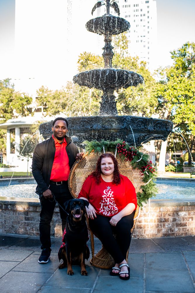 A couple poses with their black dog in front of an ornate tiered fountain in a park setting.