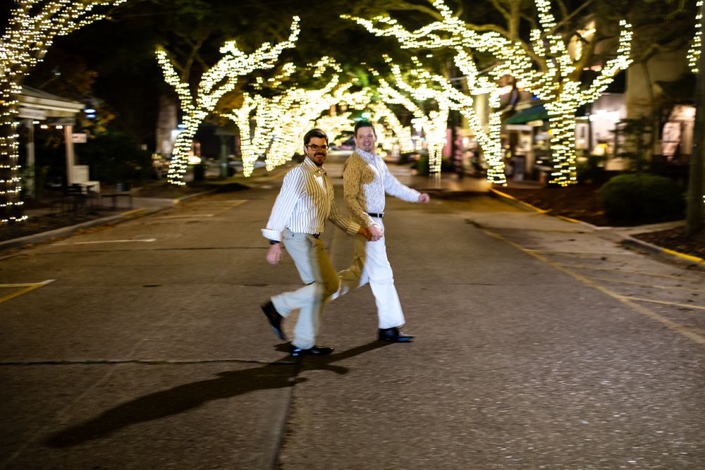 A couple strolls at night under trees illuminated with twinkling white Christmas lights along a festive street.