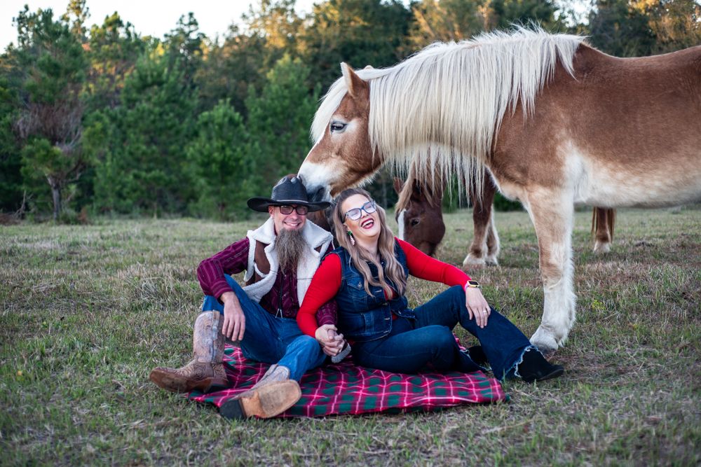 A couple relaxes on a plaid blanket in a field with their Belgian draft horse looking on.