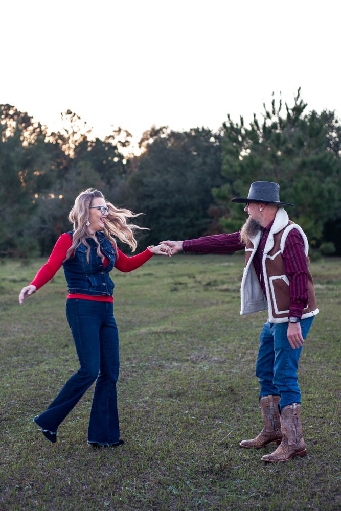 A couple in country western attire dance playfully in a grassy field at sunset.