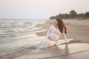 A person in a flowing white dress lies gracefully on a misty beach shoreline at sunset.