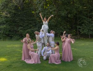 A wedding party in rose pink dresses and white outfits forms a cheerful pyramid pose on a green lawn near woods.