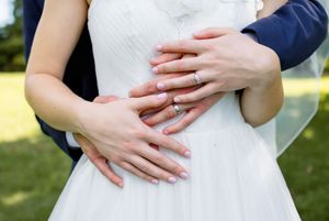 Close-up of hands with wedding rings embracing around waist in white dress outdoors.
