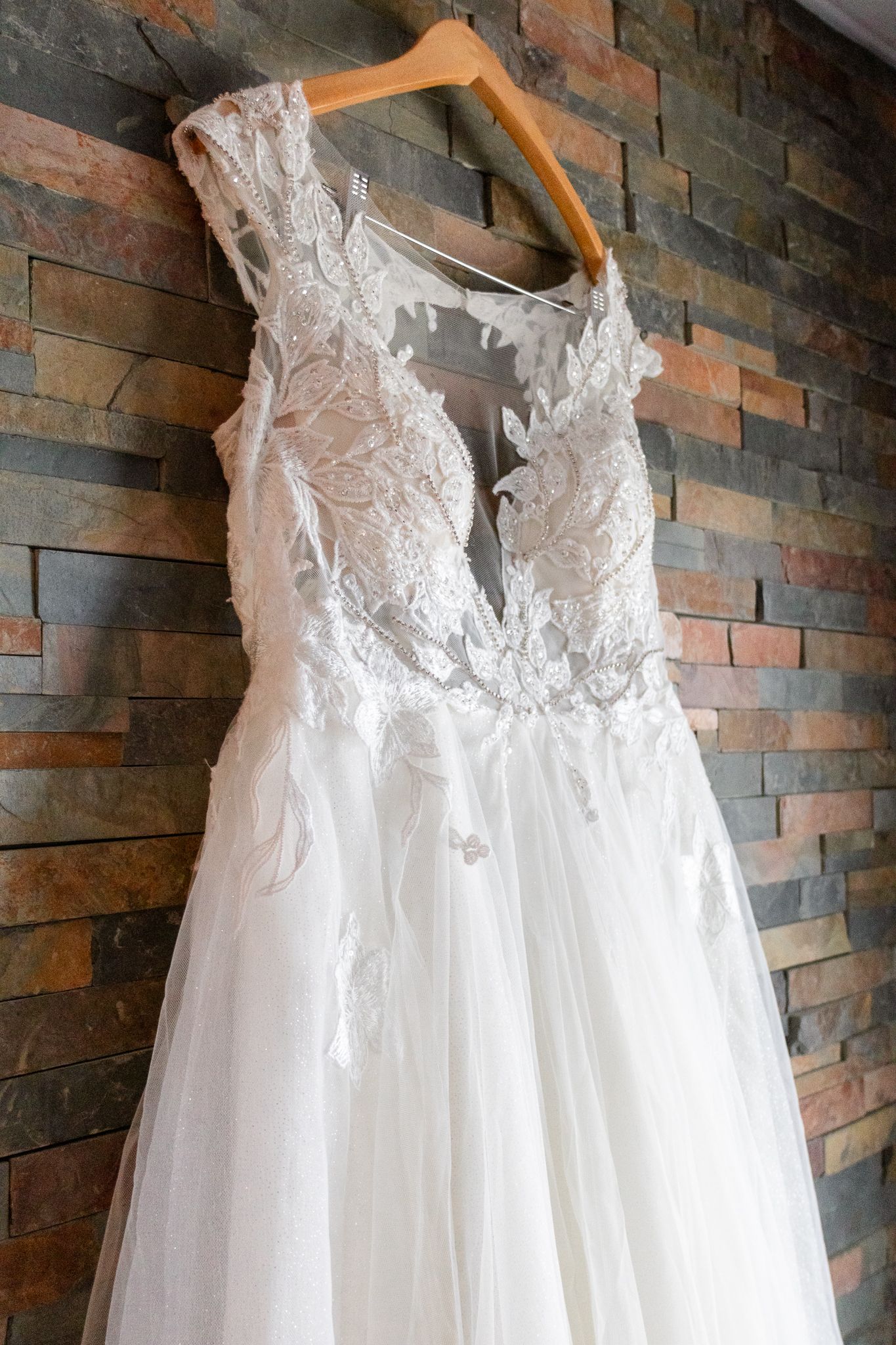 Elegant white lace wedding dress with plunging V-neckline and flowy tulle skirt hanging on wooden hanger against stone wall.