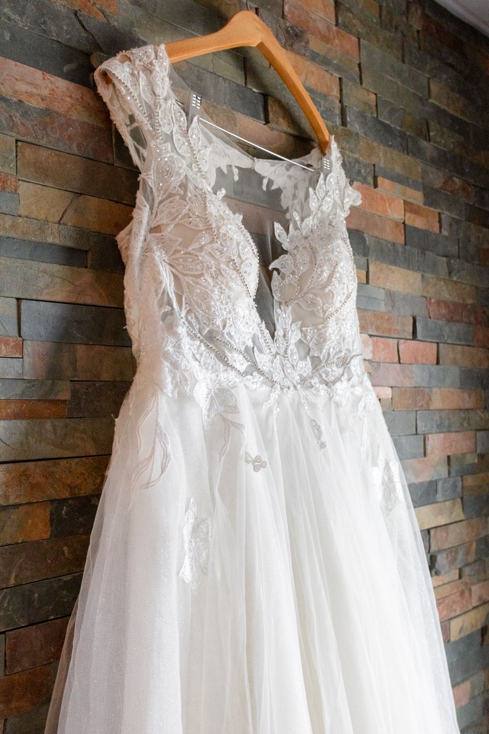 Elegant white lace wedding dress with plunging V-neckline and flowy tulle skirt hanging on wooden hanger against stone wall.