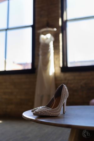 Pearl-encrusted bridal heels on a wooden table with a white wedding dress hanging in a sunlit brick loft window.