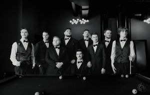 Black and white photo of nine men in formal tuxedos posing around a billiard table in an elegant pool hall.
