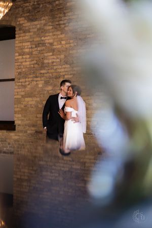 Bride and groom share a romantic moment near a brick wall, bride in white gown with veil, groom in black tuxedo.