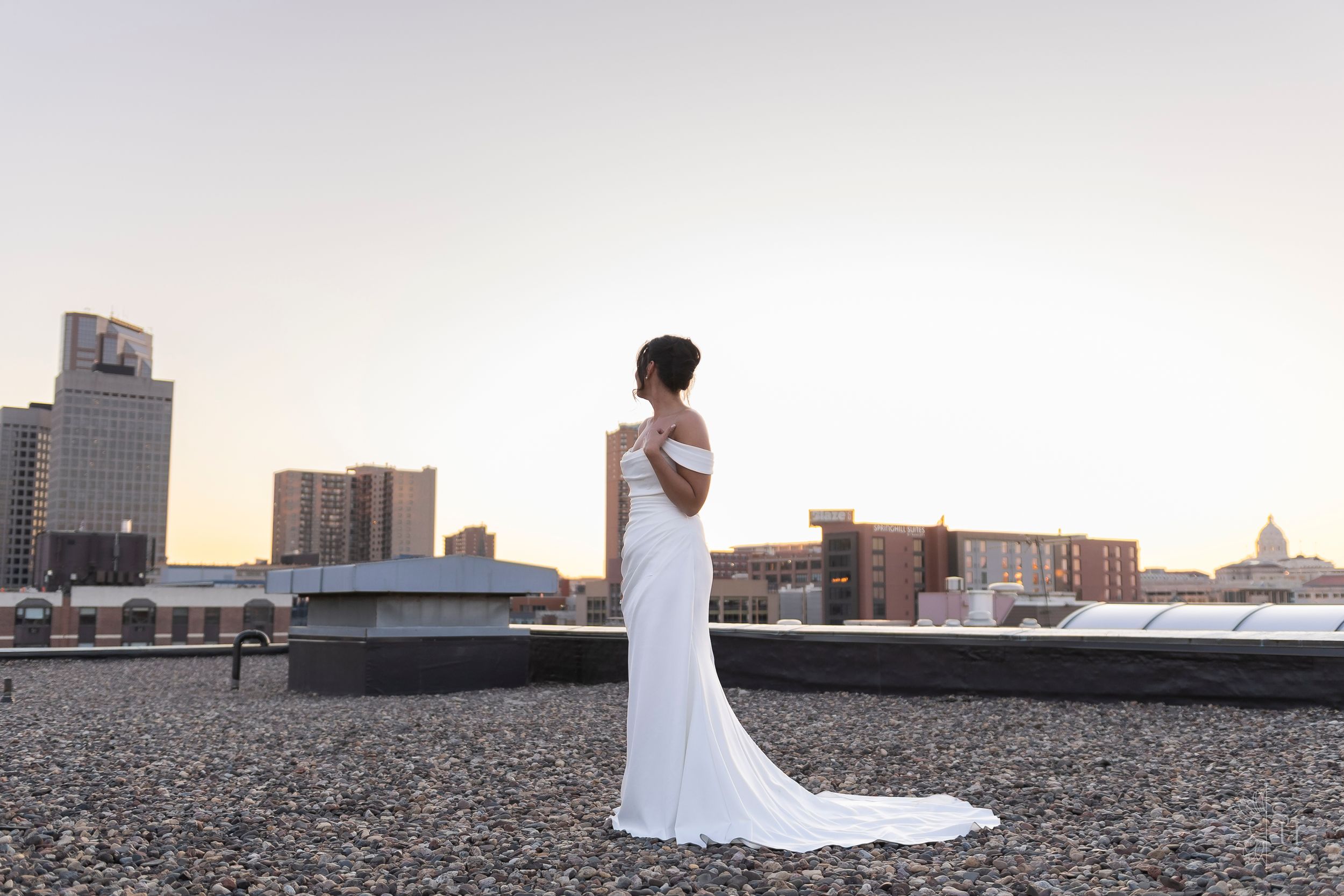 Bride in elegant white off-shoulder gown stands on rooftop with city skyline at sunset.