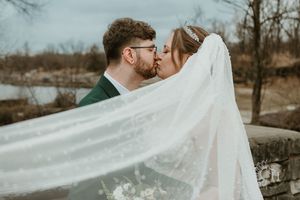 A bride and groom share a romantic kiss as her sheer white veil flows dramatically in the wind during their outdoor wedding.