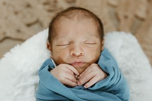 A sleeping newborn baby lies swaddled in blue fabric on soft white bedding.