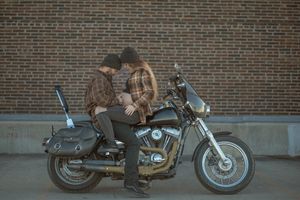 Couple sitting on parked motorcycle against brick wall at dusk.