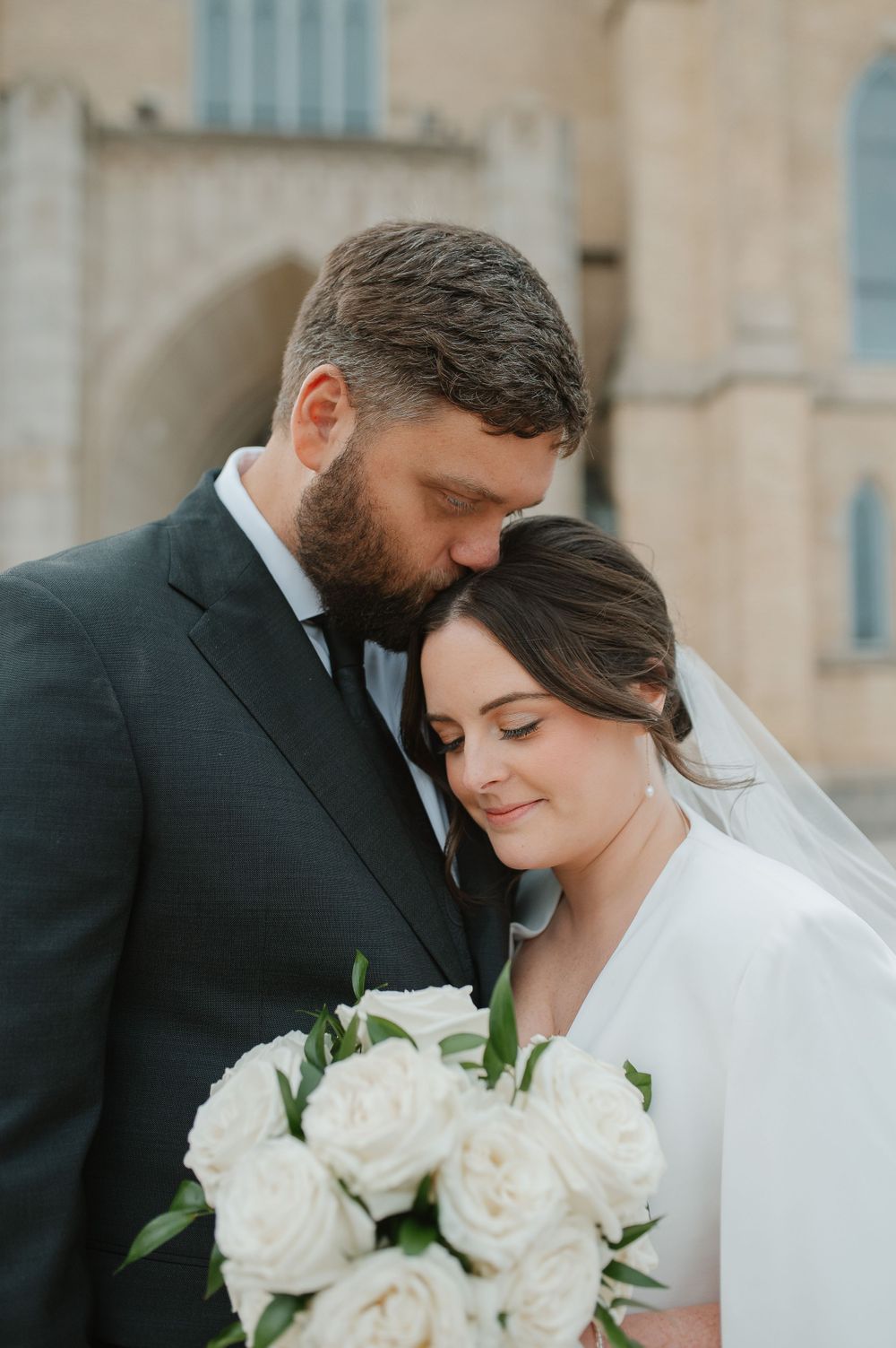 Wedding couple shares intimate moment against stone building with white rose bouquet.