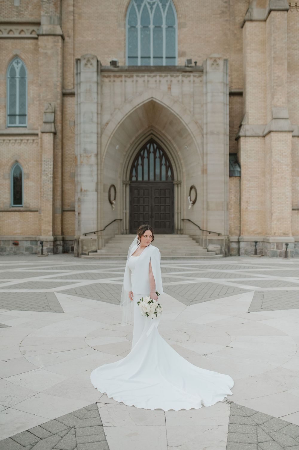 Elegant bride in flowing white dress poses in front of gothic church architecture.