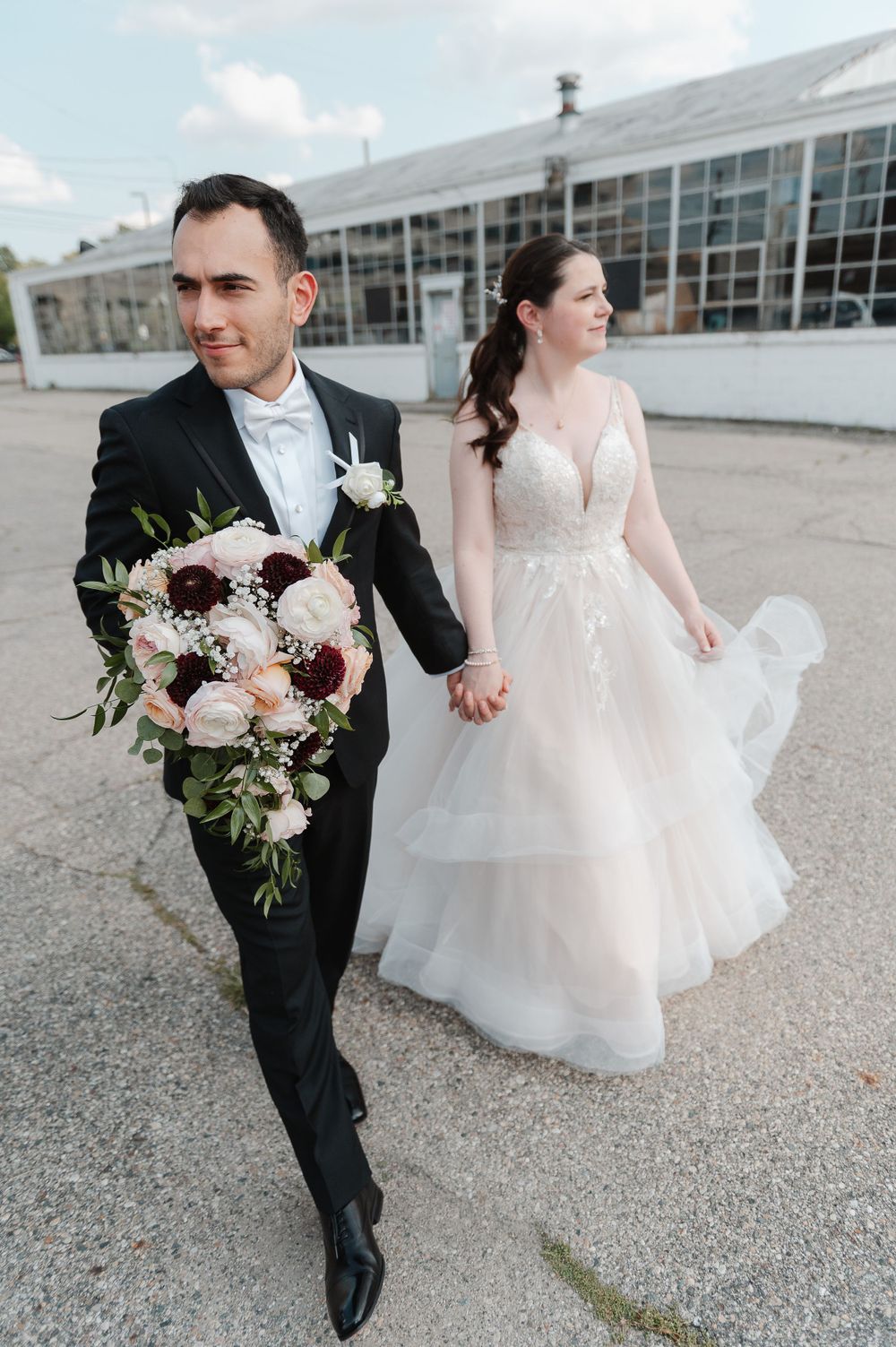Wedding couple walking outdoors with pink and white bouquet near modern building.
