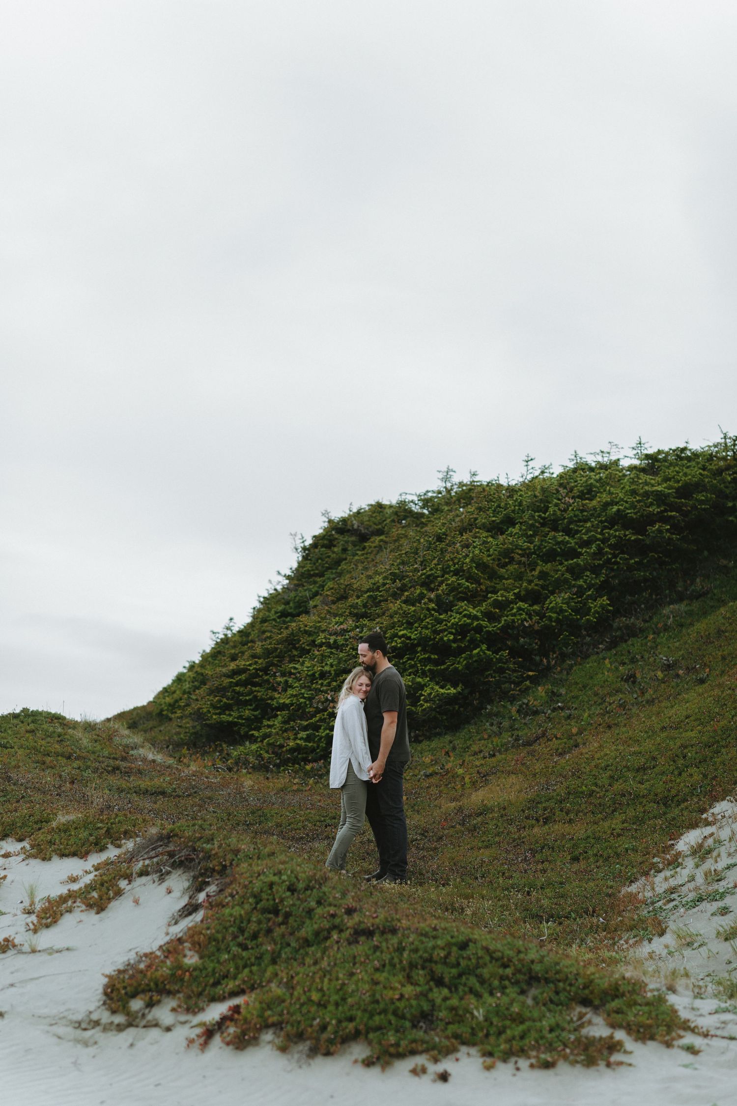 A couple stands close together on a coastal hill with green vegetation against an overcast sky and sandy beach.