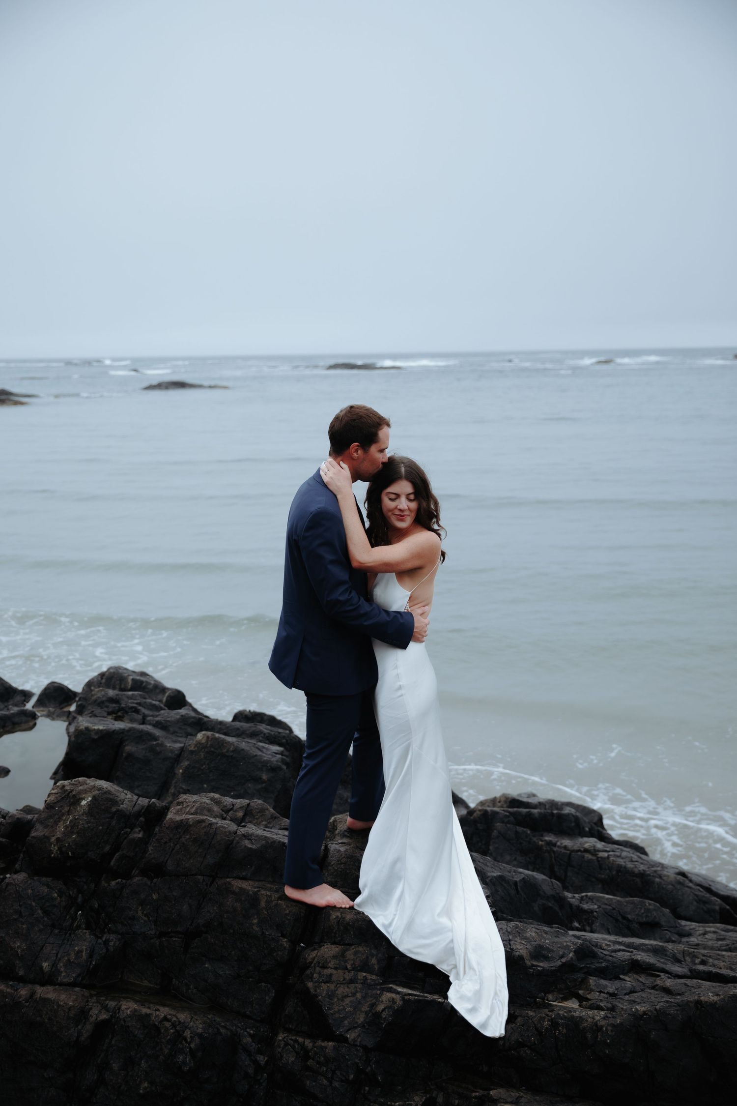 A couple shares a romantic embrace on rocky coastal cliffs overlooking a moody gray ocean.