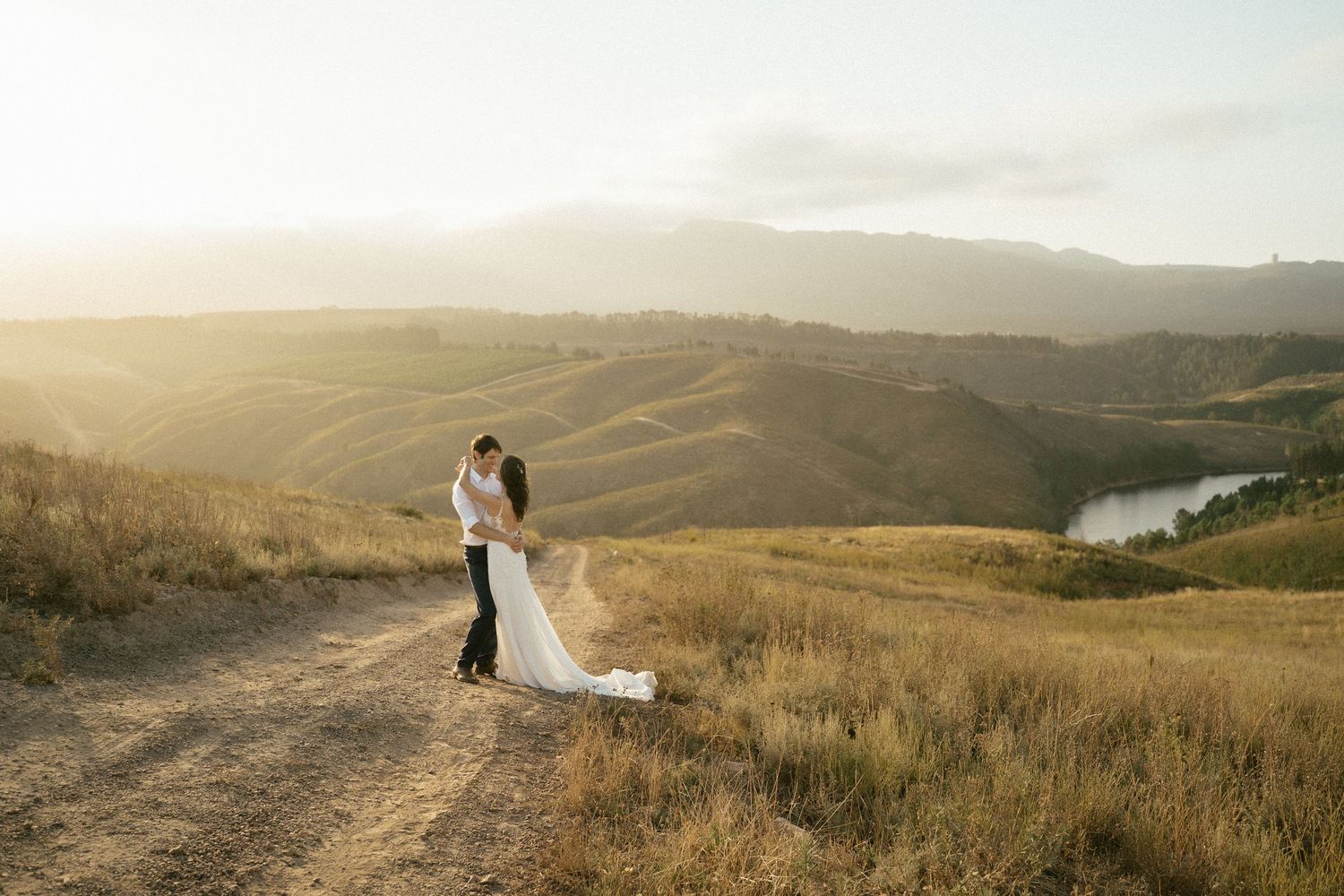 Newlyweds embrace on a hilltop during golden hour overlooking rolling hills and a winding river.