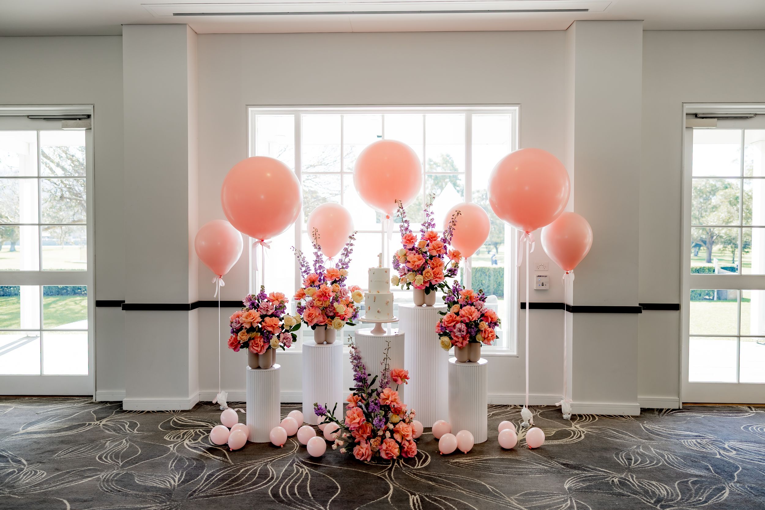 Modern event space with pink balloons and floral arrangements decorating a bright white room with large windows.