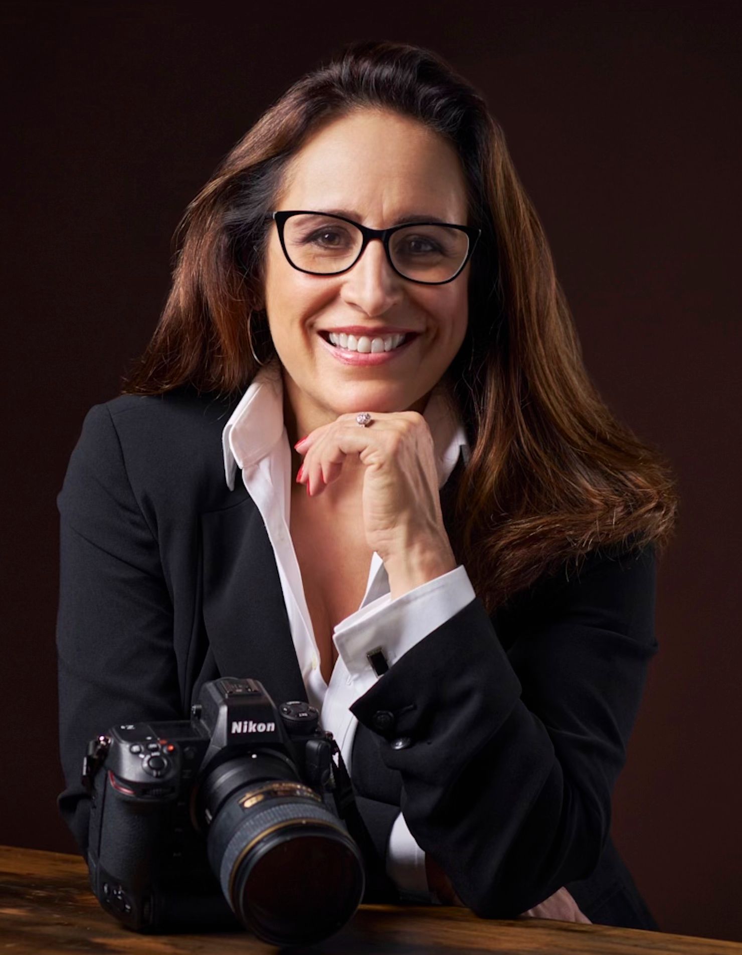 Professional female photographer smiling confidently with a Nikon camera, wearing glasses and a black blazer.