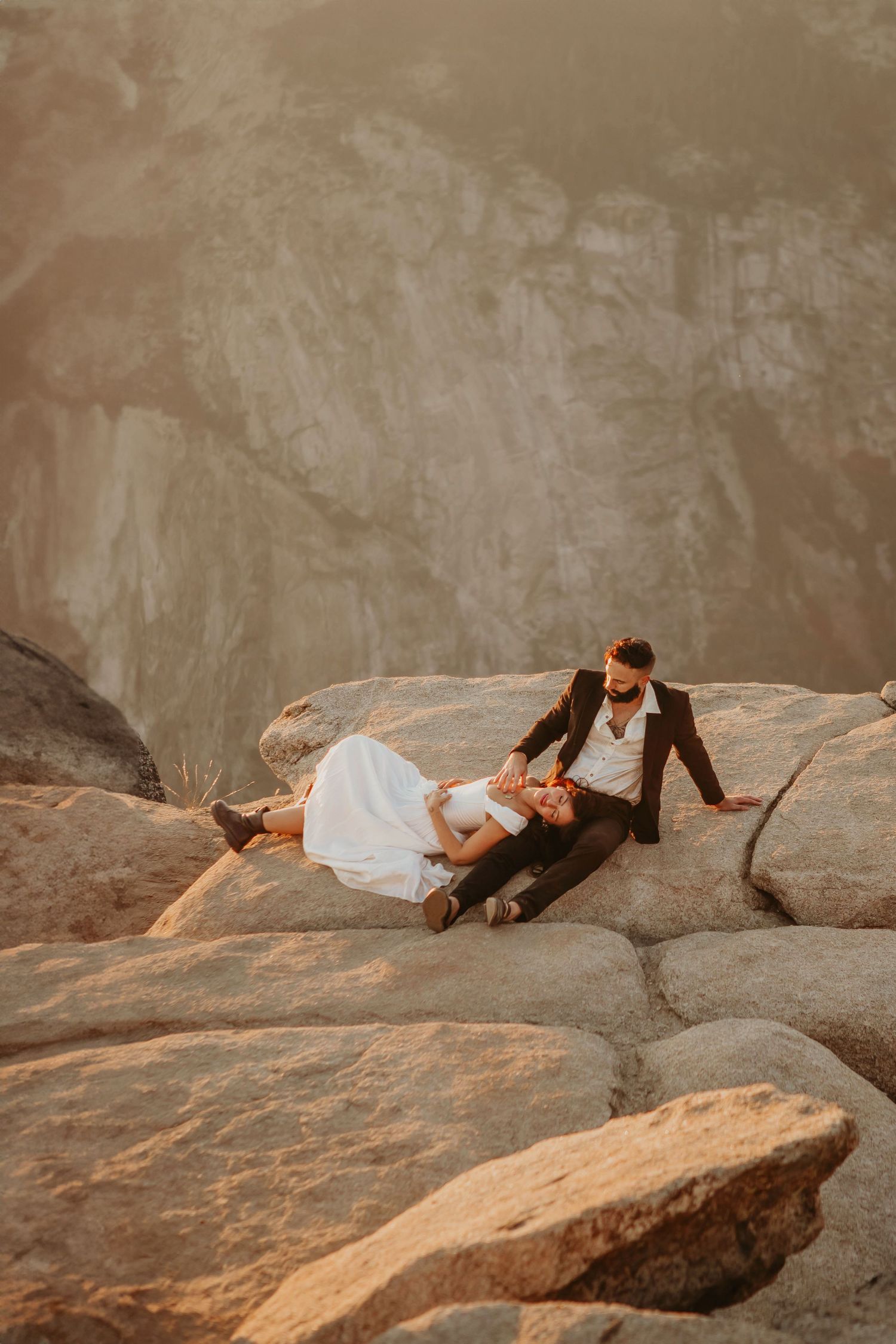 Couple in wedding attire relaxing on rocky mountain ledge at sunset.