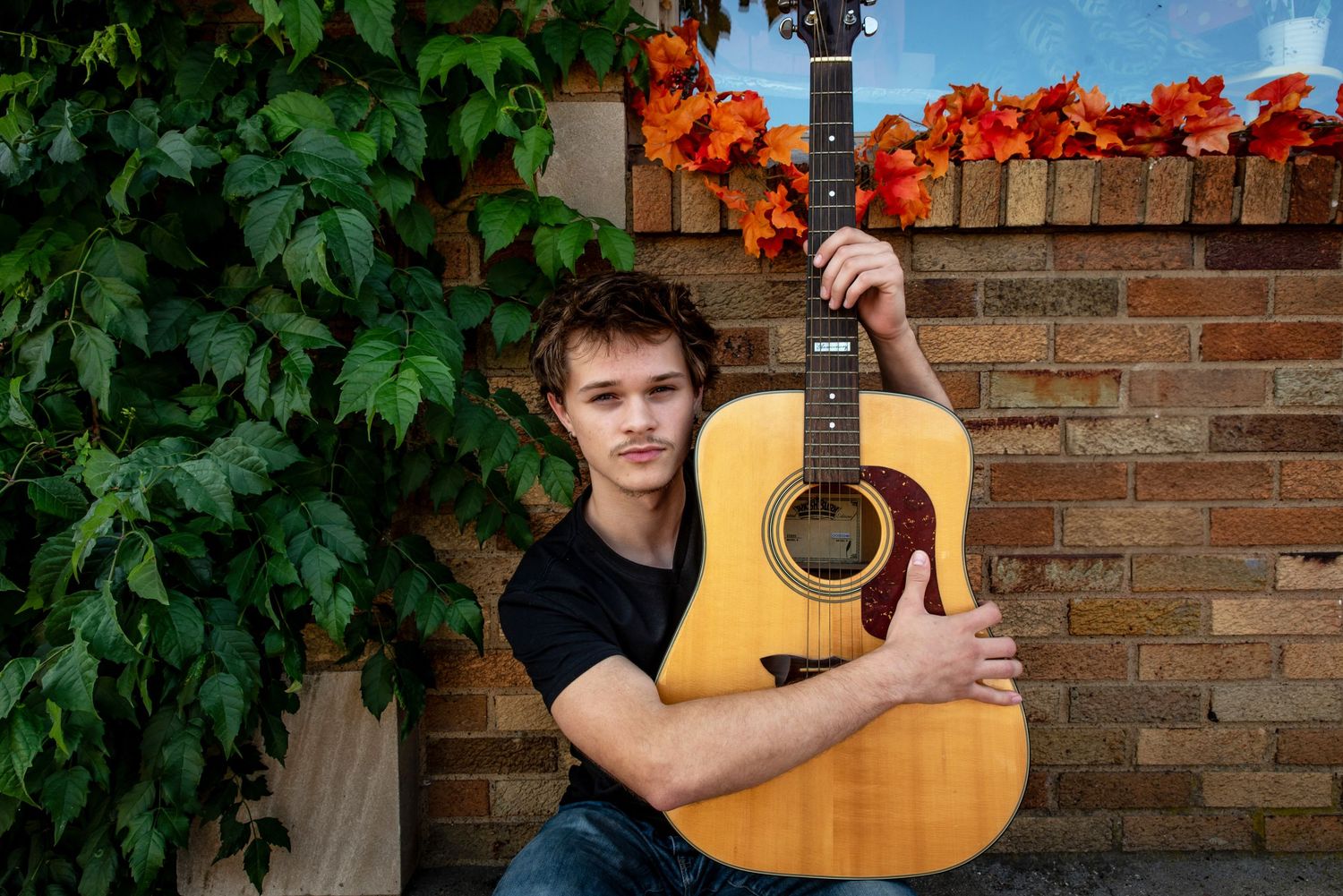 Portrait with acoustic guitar against brick wall decorated with orange lights and greenery.