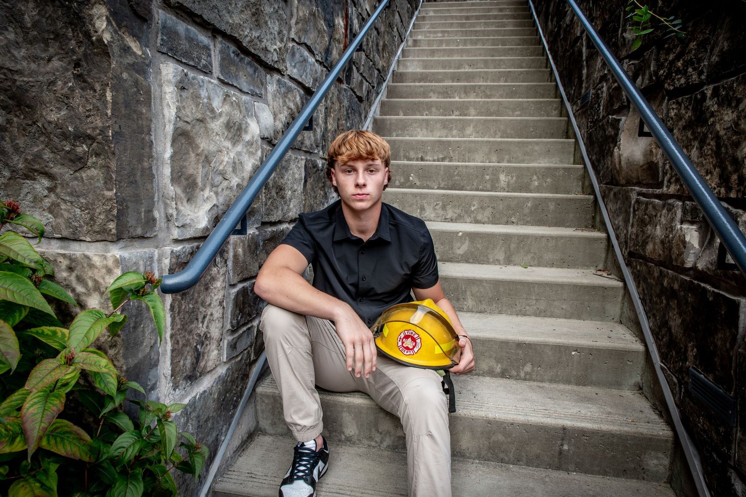 Subject sitting on concrete steps holding a yellow hard hat in an outdoor setting.