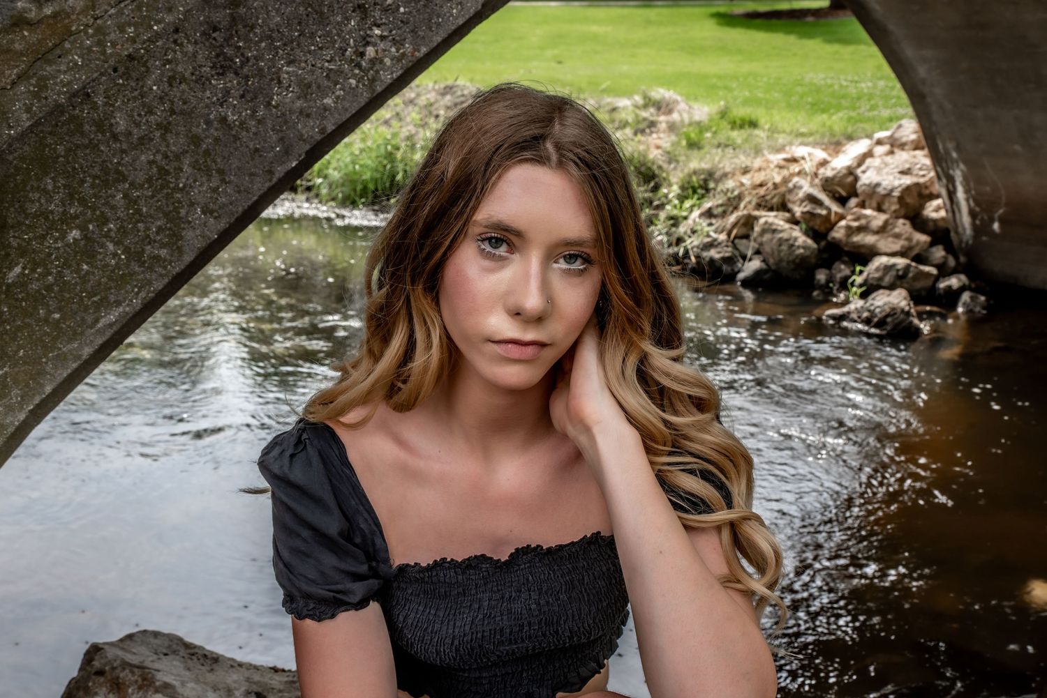 A model in a black dress poses beneath a bridge near a stream with rocks and greenery in the background.