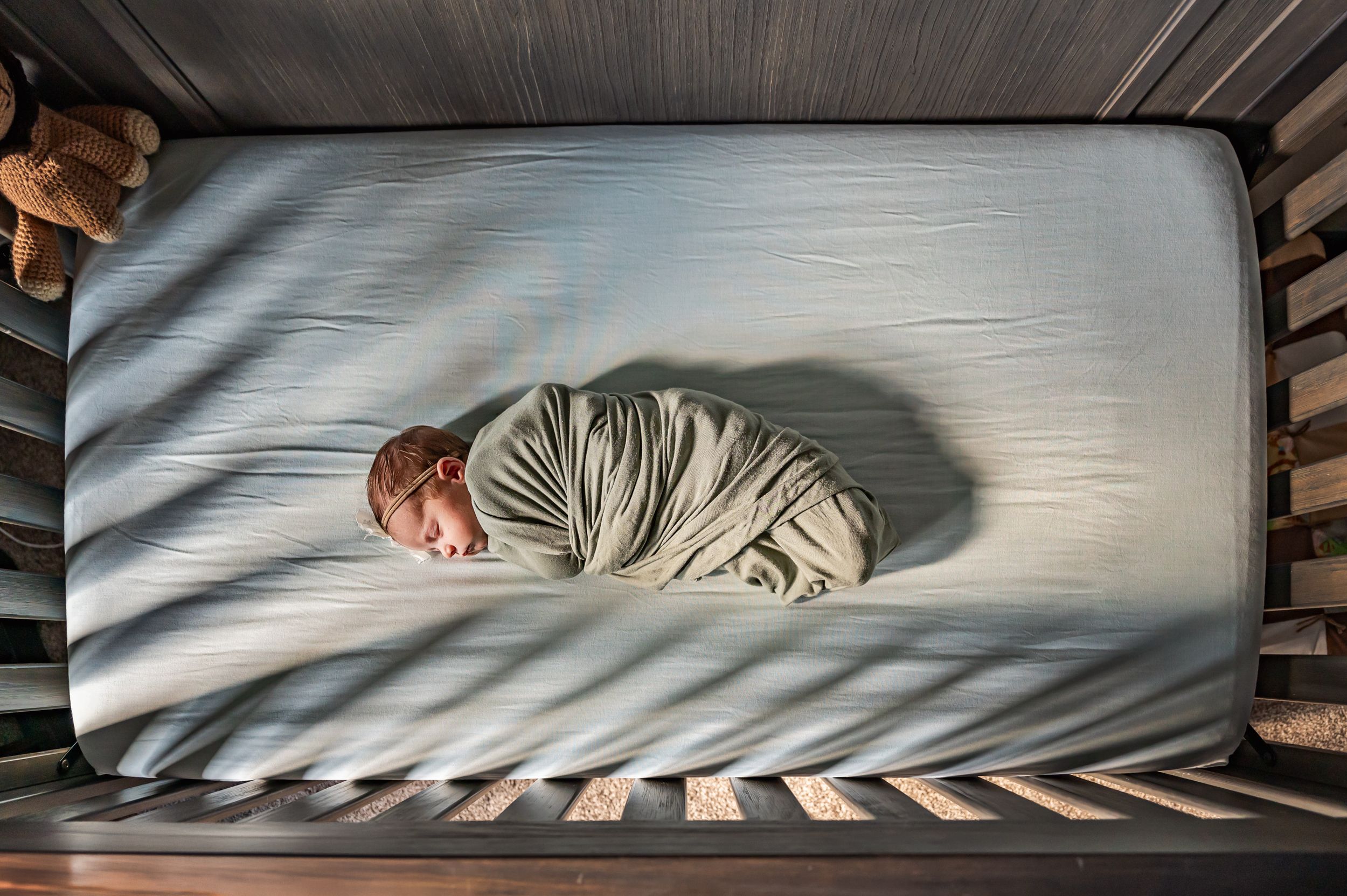 A swaddled newborn sleeps peacefully in a wooden crib viewed from above.