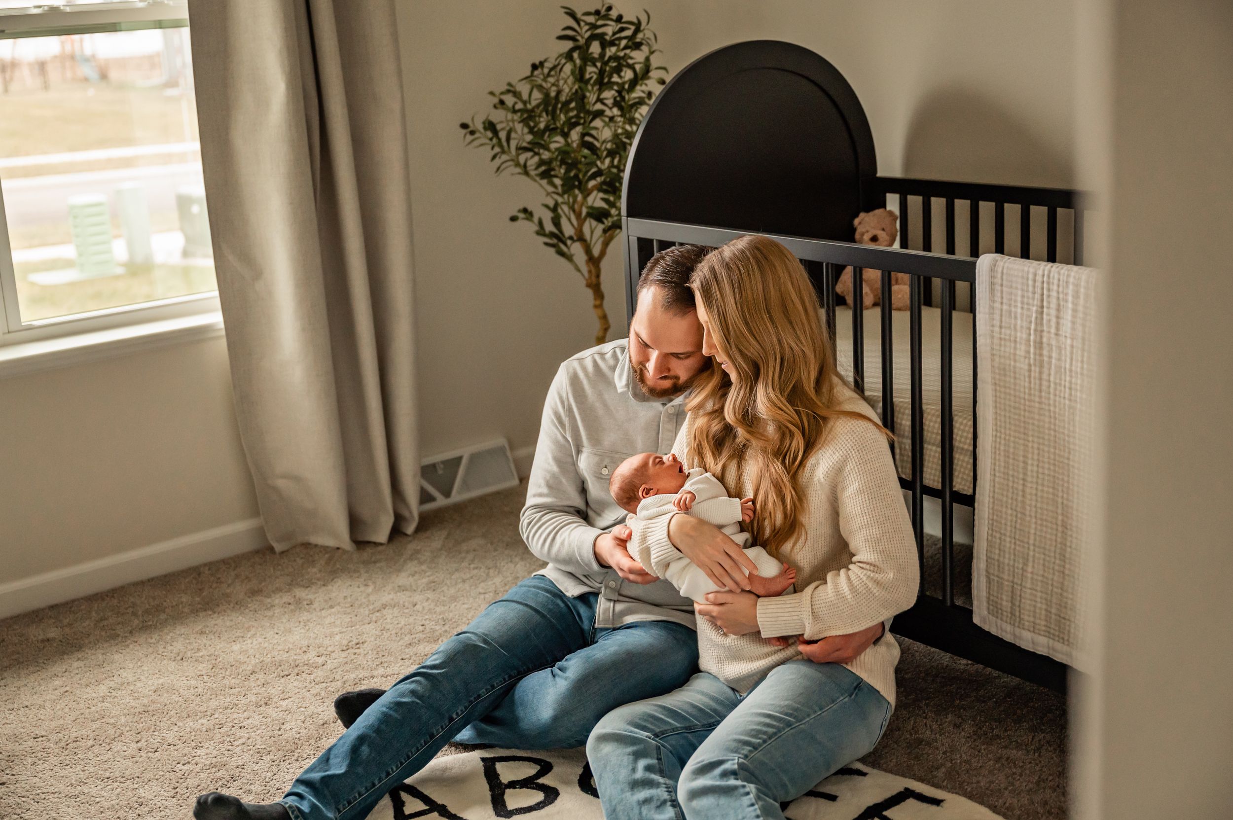 A couple sits on a carpeted floor beside a black crib, sharing a tender moment.