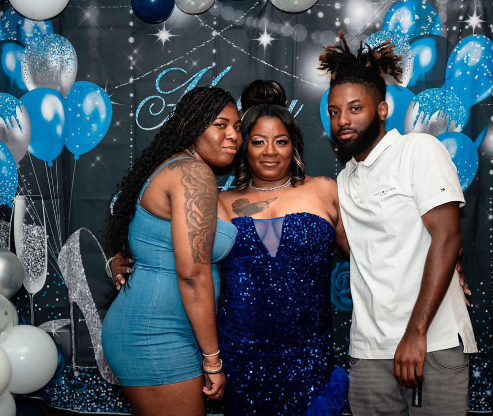 Three people pose together at a party with blue and white balloons in the background.