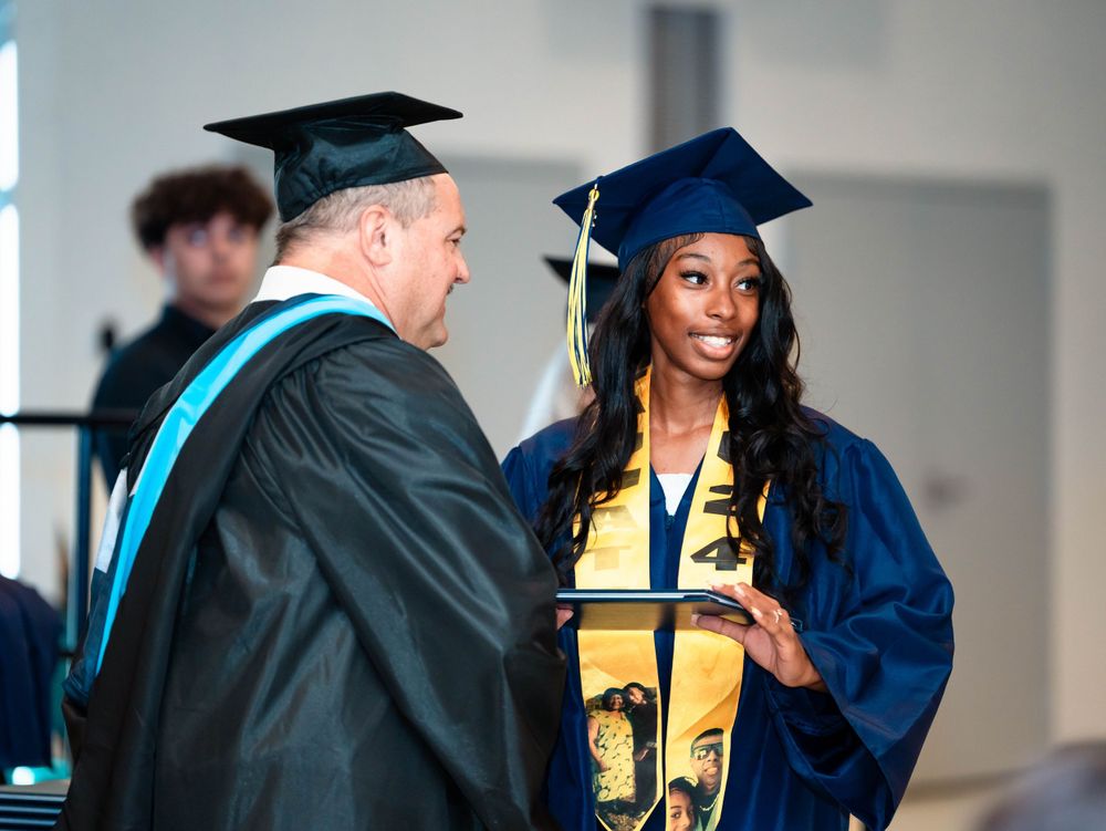 A graduation ceremony moment with a graduate in blue cap and gown receiving recognition.