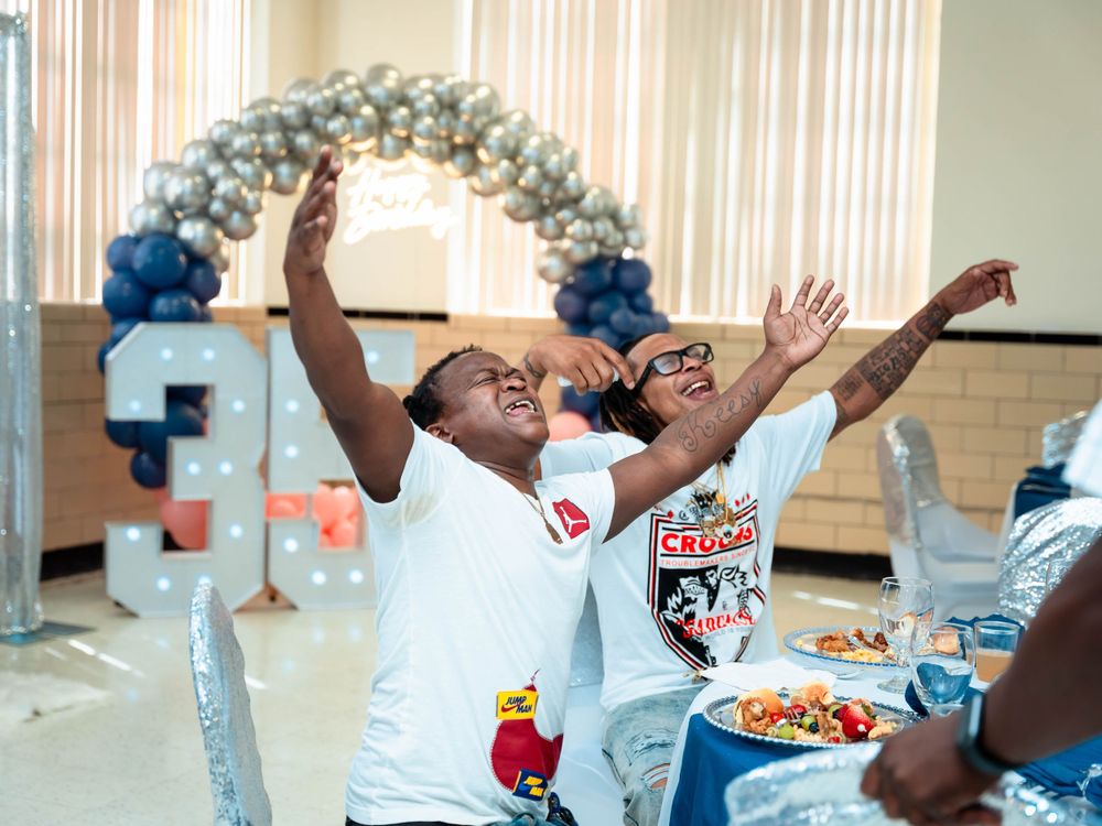 A person in white t-shirt celebrates enthusiastically under a balloon arch at an indoor event.