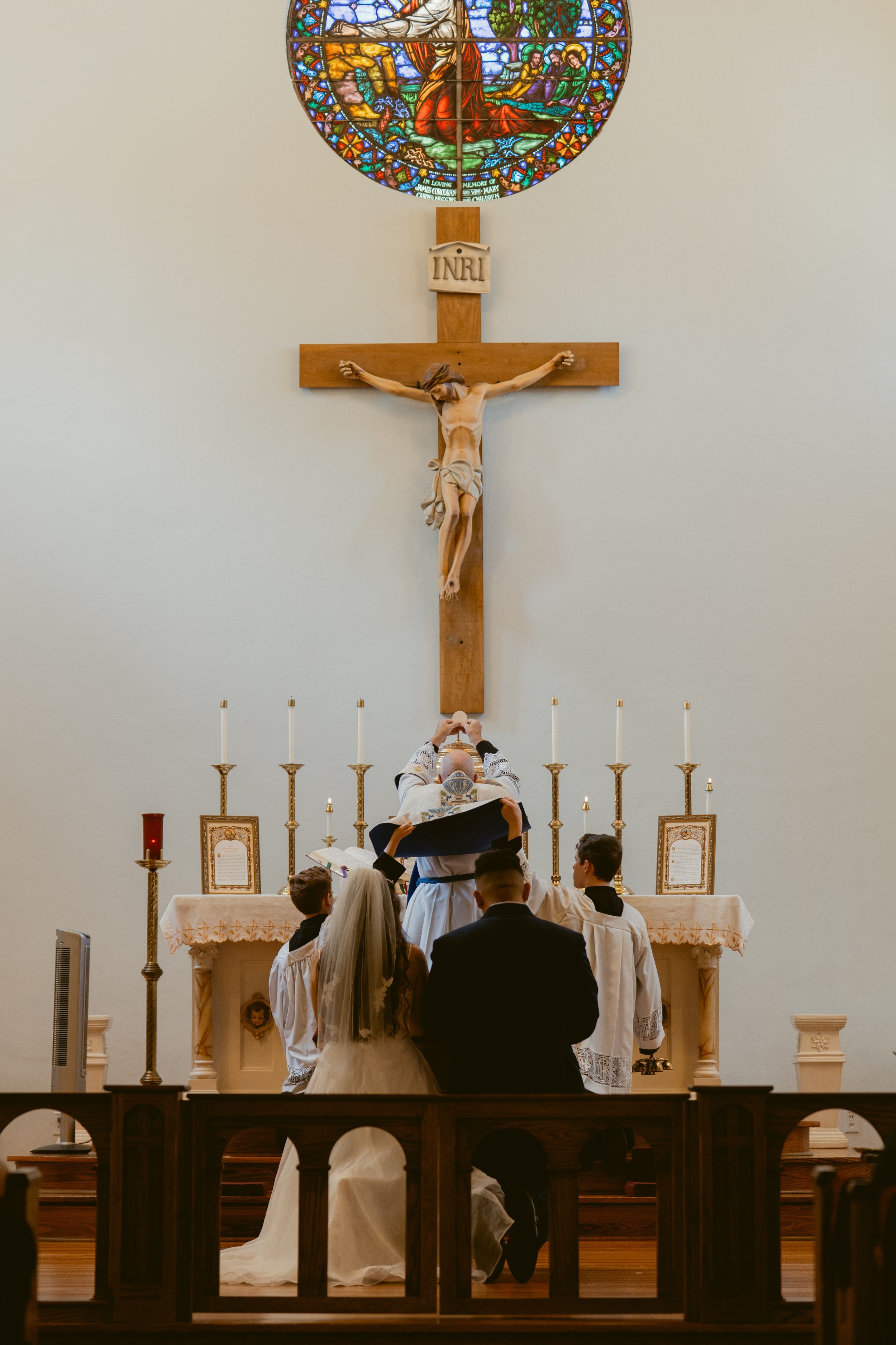Catholic wedding ceremony at church altar with crucifix, stained glass window, candles, and couple kneeling before priest.
