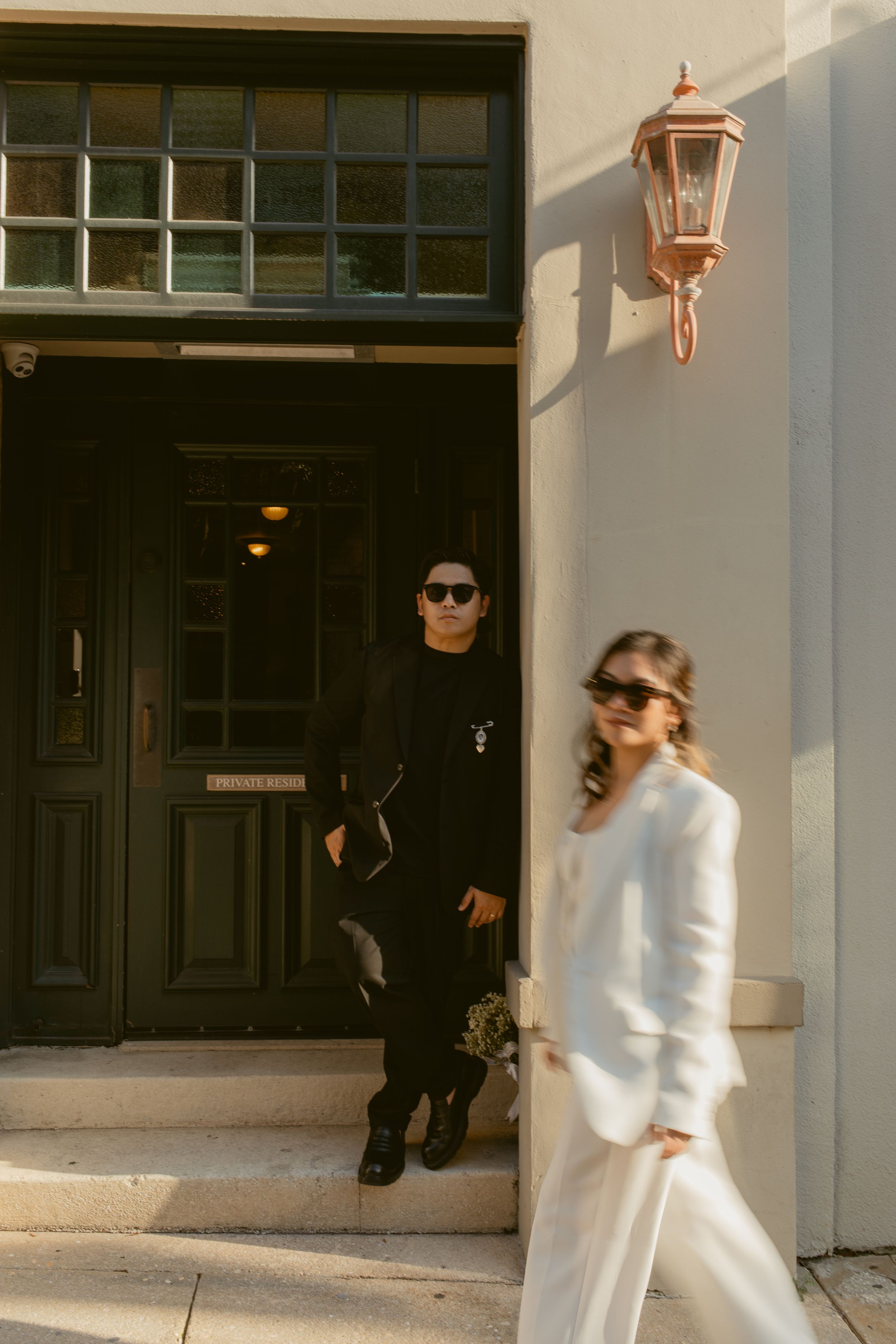 A couple in formal wedding attire embraces outside a building with white walls and dark doors.