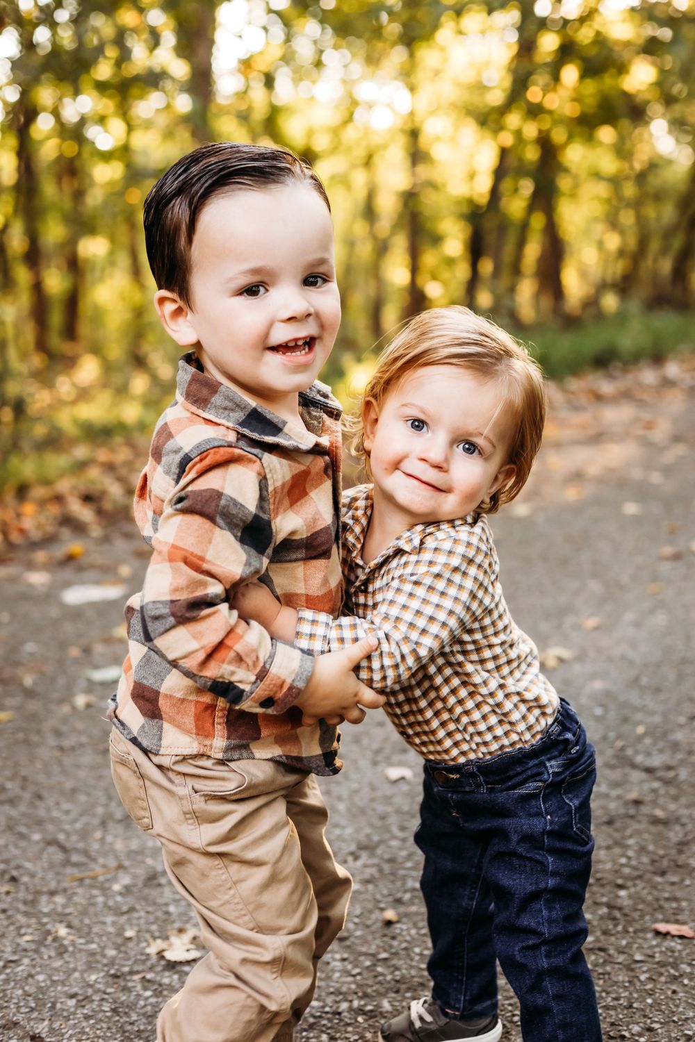 Two young children in plaid and checkered outfits share playful hugs on forest path New Haven, CT.
