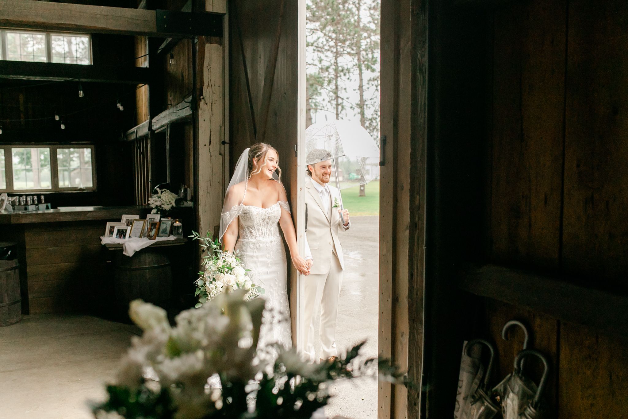 Bride in a white gown and her partner in a light suit stand together in a rustic barn doorway with white floral arrangements in front.