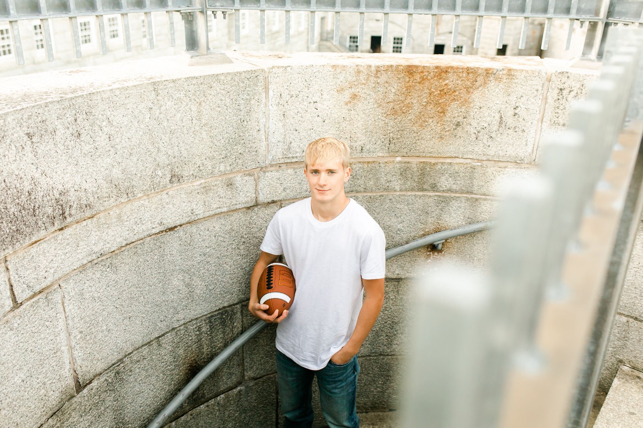Teen boy in a white t-shirt and jeans holds a football while standing in a curved concrete stadium stairwell for a senior portrait.