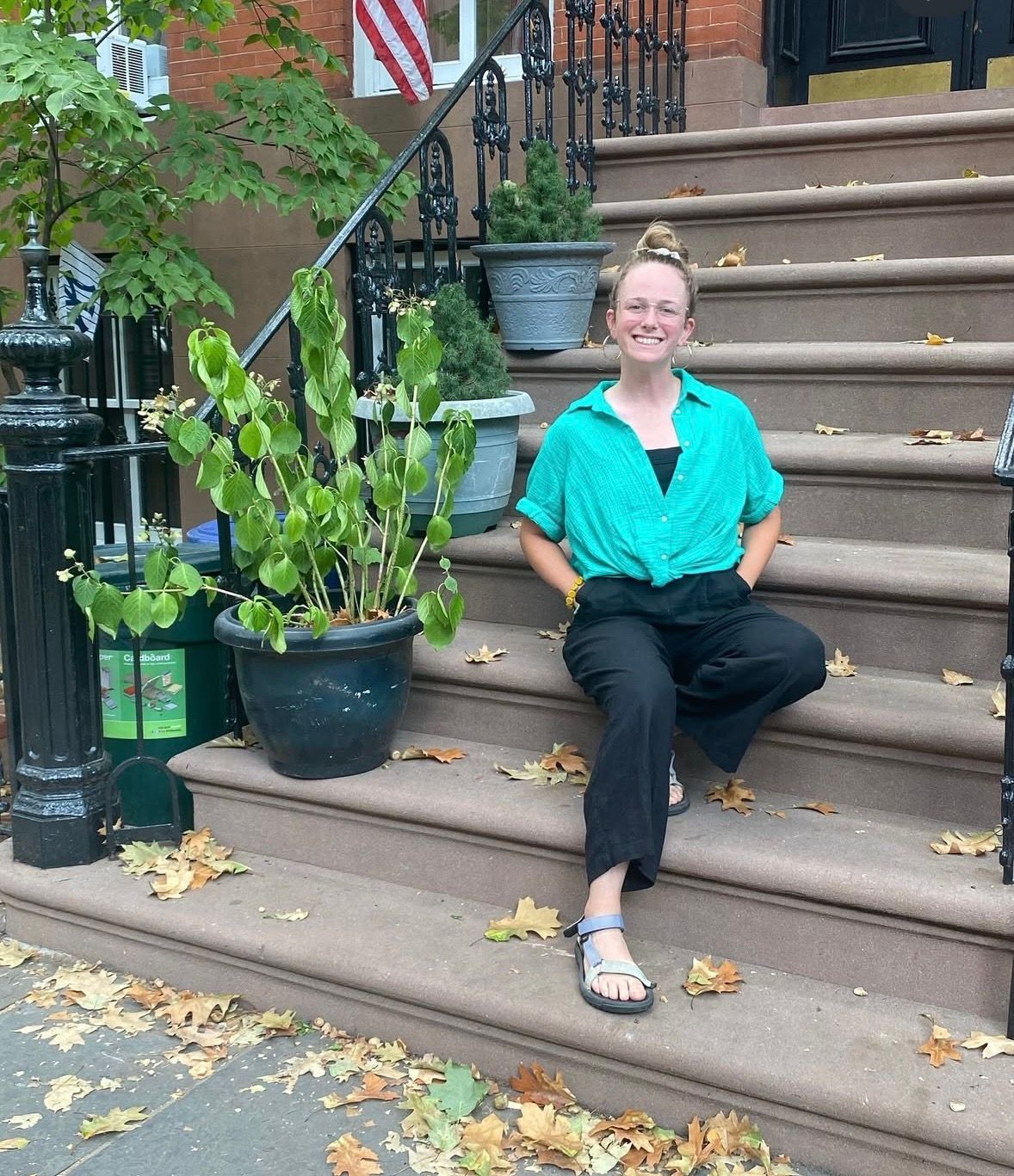 Woman in teal shirt and black pants sitting on brownstone steps beside potted plants on a city street.