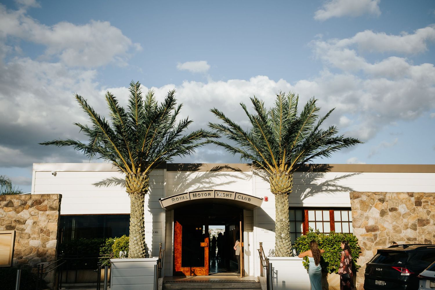 Elegant venue entrance with two tall palm trees flanking white building under blue cloudy sky.