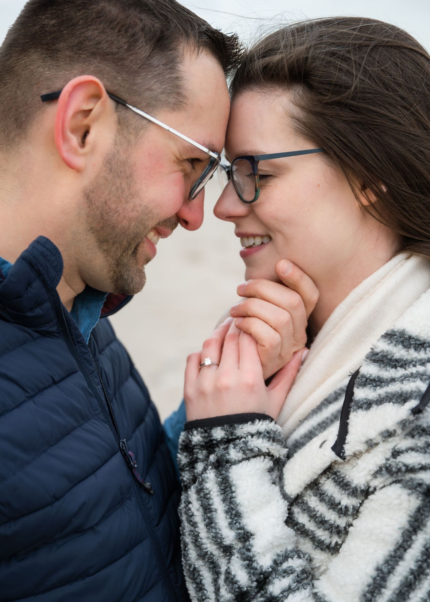 A smiling couple in glasses touch foreheads tenderly, holding hands outdoors, wearing winter jackets in a romantic moment.