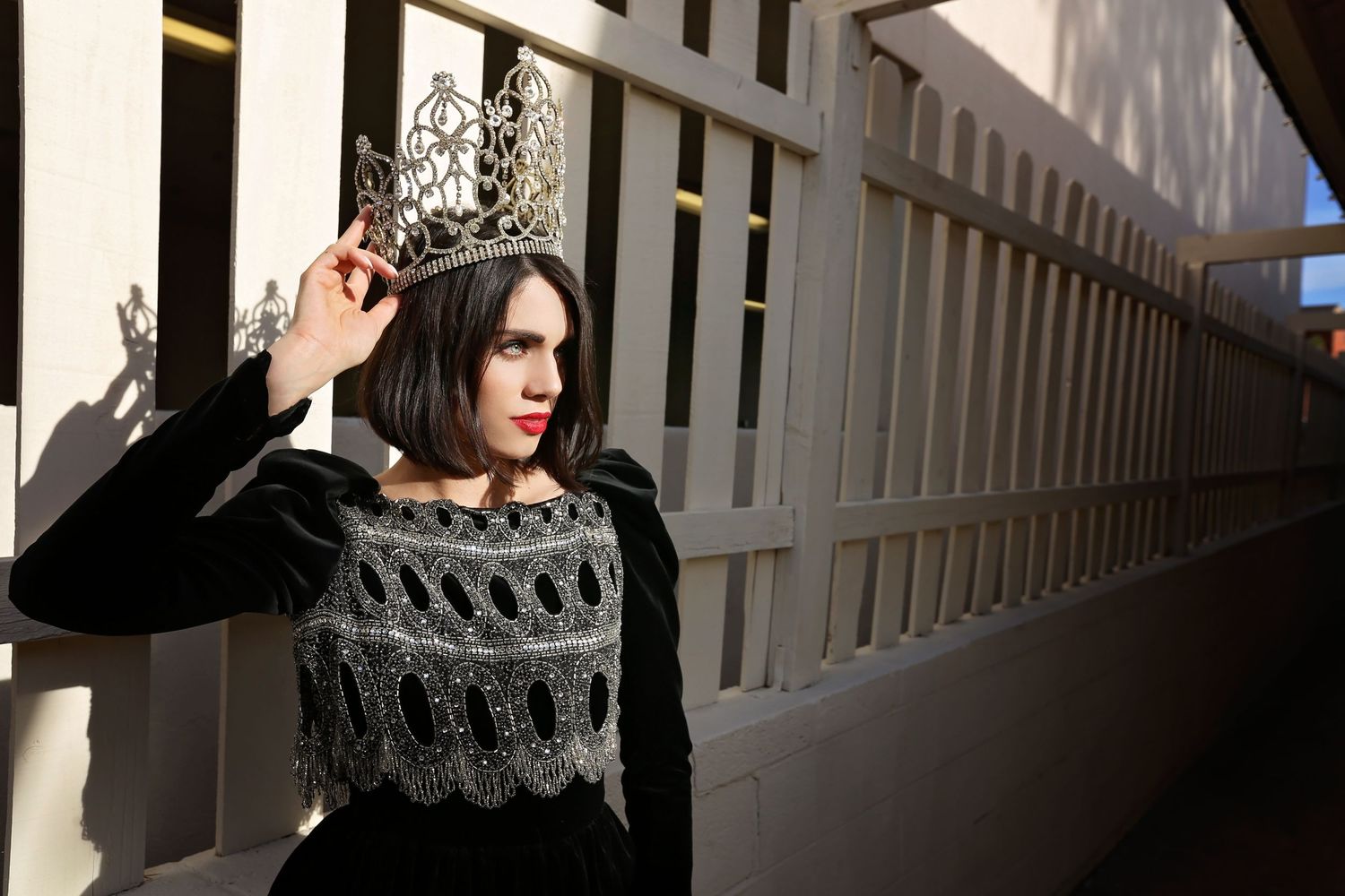 Woman in a black velvet embroidered dress and red lipstick lifts a sparkling tiara onto her head beside a white slatted fence.