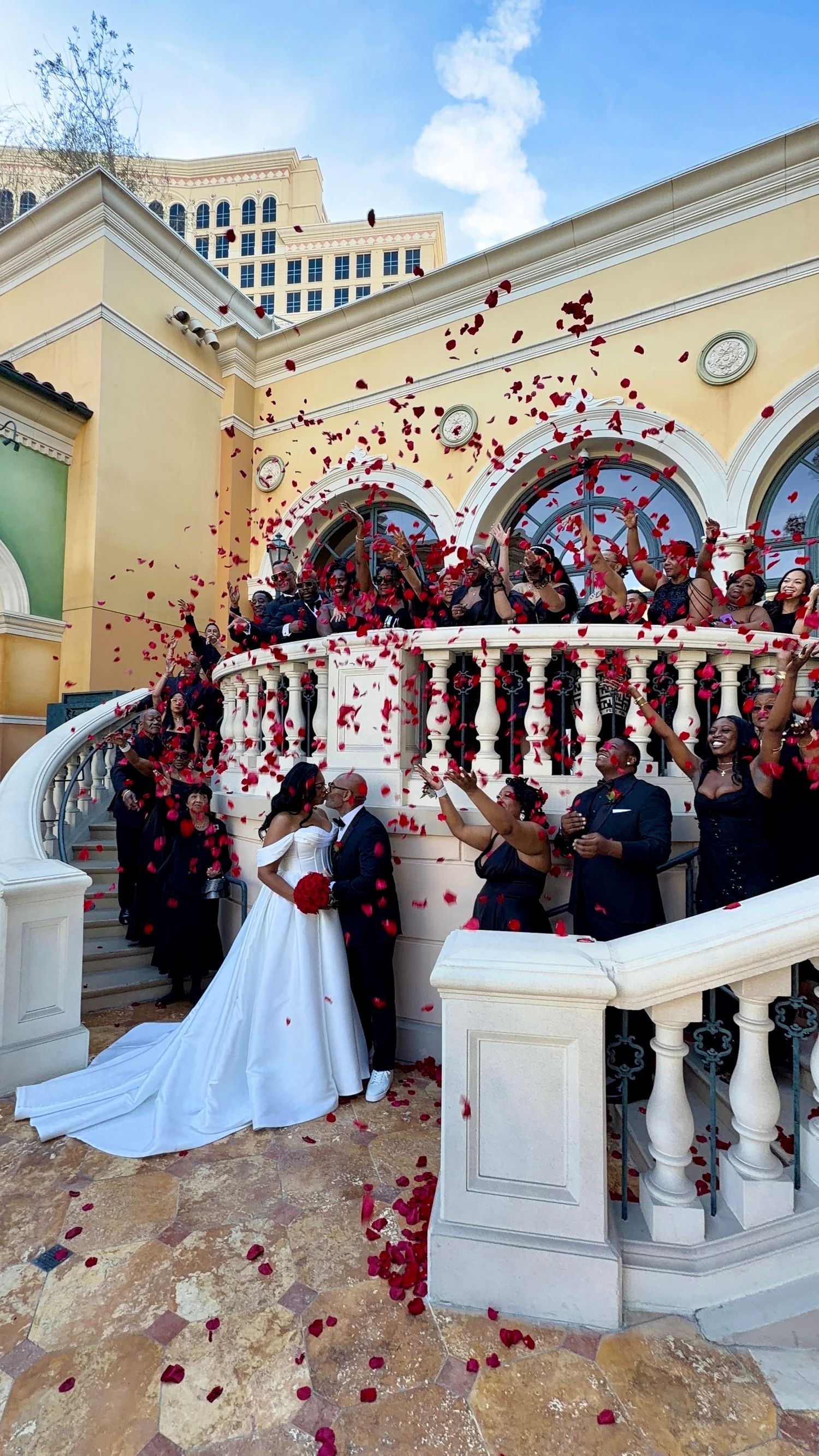 Bride and groom kiss on elegant staircase as wedding party throws red rose petals at a Las Vegas venue.