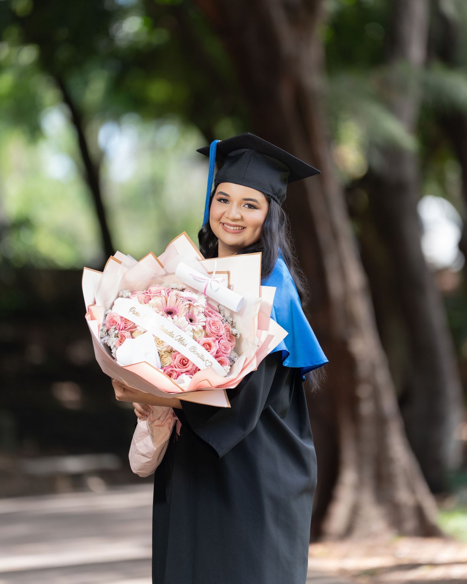 Smiling graduate in black cap and gown sits holding diploma folder, celebrating academic achievement.