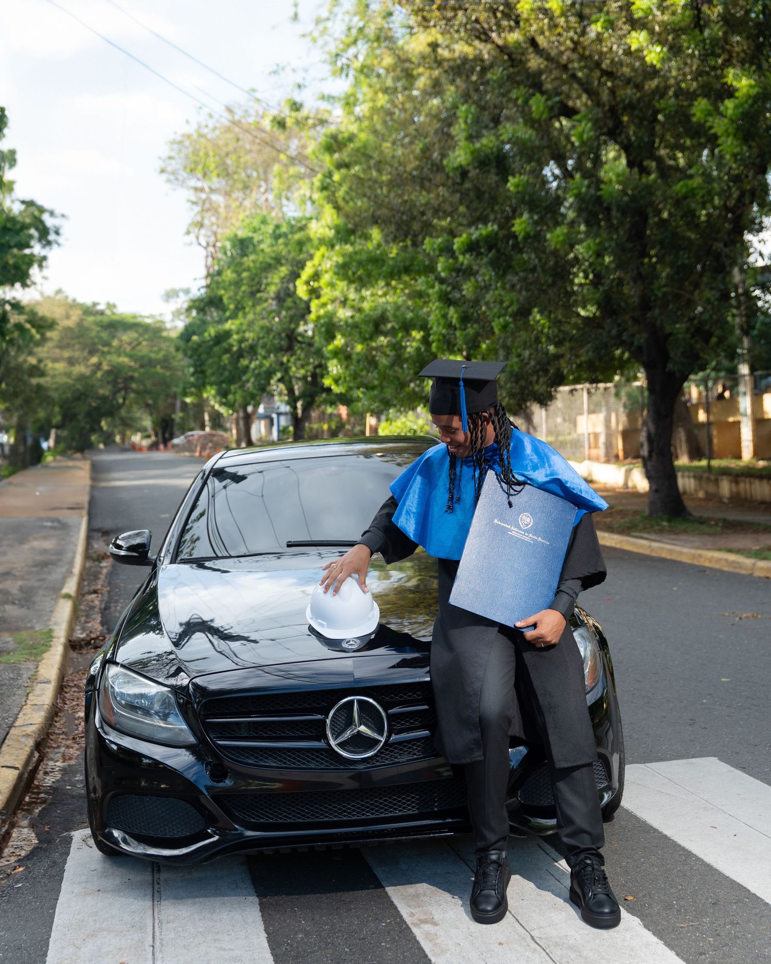 Smiling graduate in black cap and gown sits holding diploma folder, celebrating academic achievement.