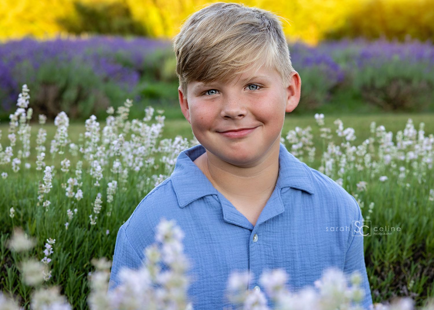 Smiling blond boy in blue shirt posing in a colorful lavender and wildflower field outdoors.